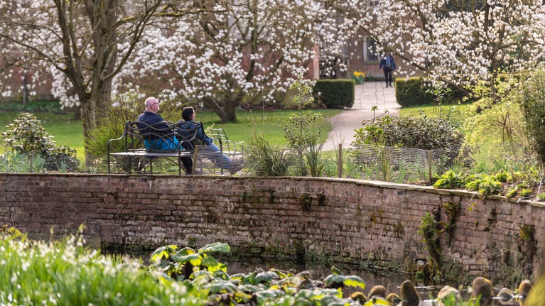 A man and a woman sat on a bench facing away from the camera. They are sat underneath white blossom trees. The red brick Georgian house of Dunham Massey can be seen in the background. A brick canal wall surrounded by green flowers can be seen in the foreground.